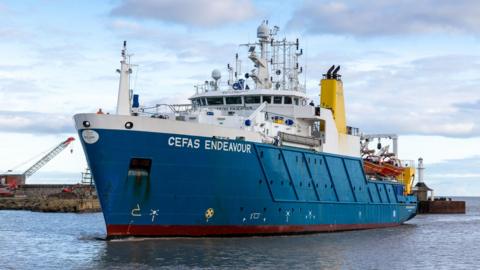 The research vessel Cefas Endeavour coming in to harbour. It has a blue hull and white wheelhouse. On the wheelhouse roof are many piece of radar and satellite technology. Lifeboats can be seen towards the stern. The sea is calm and there is a quay behind the boat.