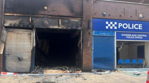 A burned out building, with a gaping shopfront and metal door shutter all damaged by fire, next to a building with a blue shutter and with blue signage reading  "Sunderland Central Police Office", with the glass frontage smashed