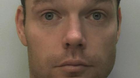 A mugshot of Daniel Lynch. He is raising his eyebrows and his eyes are wide open. He has short dark brown hair and is unshaven. He is wearing a pale brown sweatshirt.