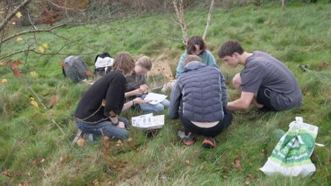 Five students crouching in a meadow looking at a soil sample and counting the number of different species of worms. They had an identification sheet of worms found in the UK.