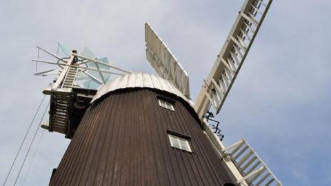 Wicken windmill in Wicken near Sohan in Cambridgeshire. The windmill has a brown exterior, reaching high into the sky. The picture has been taken from the ground and the white sails of the mill are stationery. The picture has been taken on a day with a blue sky and some overcast cloud. 