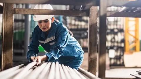 Woman in blue overalls and  white hard hat placing metal poles in a pile 