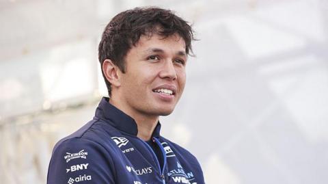 Alex Albon looking into the distance past the camera in a closely cropped photo. He has short wavy dark hair and is wearing a navy blue Williams team zipped jacket. The photo is taken at the Japanese Grand Prix but the background behind him is just white.