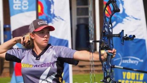 A woman wearing a had and a sports polo top holding  a compound bow up high. Blue archery banners are in the background.
