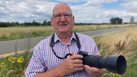 David Fyfe, wearing glasses and checked shirt, holding a black long-lens camera. He is smiling as he stands on an overgrown grass verge beside a road under a cloudy blue sky.