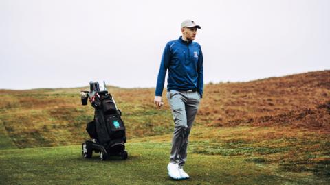 Golfer Josh Simpson on the fairway of a golf course with his buggy behind him.