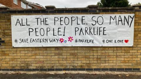 A brick wall, on which is attached a white banner saying "All the people, so many people! Parklife." A hashtag below says "Save Eastern Way"