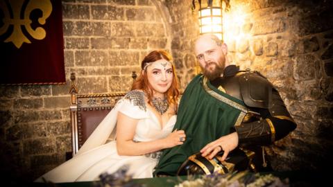 Seated portrait of two people in medieval-inspired attire inside a stone-walled room lit by a warm lantern. Jasmine who has ginger hair wears a white flowing dress with decorative silver shoulder pieces and a jewelled headpiece, while her husband wears dark armour with a green sash draped across the chest. They sit closely together on an ornate wooden chair beside a red banner with a gold emblem.