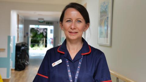 A woman wearing a nurses uniform with the name pin Claire on it standing in the hallway of a hospital. The back door is open and grassland and trees can be seen.