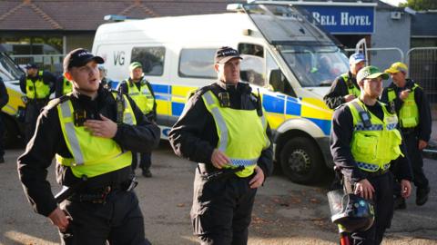 A group of police officers in fluorescent tabards speaking into talkback devices and holding riot helmets - there is a large police van in the background at the entrance to a building with a Bell Hotel sign 