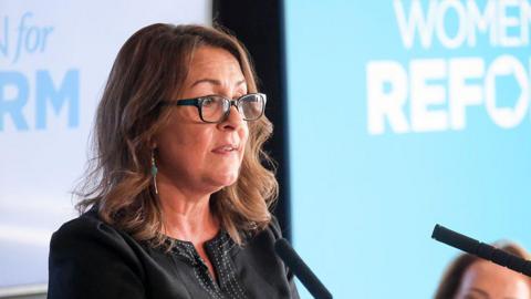 Linden Kemkaran wearing glasses and talking into two podium microphones. Light blue and white backgrounds can be seen behind her.