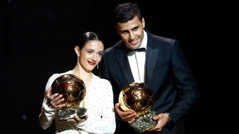 Ballon d'Or winners from 2024 Aitana Bonmatí and Rodri holding their trophies stood side by side at the awards ceremony