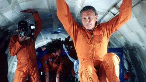 Men gather in a white space, all are wearing orange jumpsuits and hold the roof.