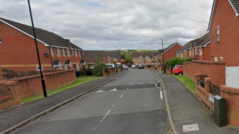 A wide image of Wolston Way in Coventry. Red brick homes line the street with cars parked in front of properties. There aer some grass verges visible with trees in the distance