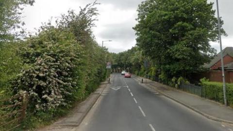 A road view of Chew Moor Lane in Bolton with two cars approaching from a distance, with tall trees either side of the highway. 