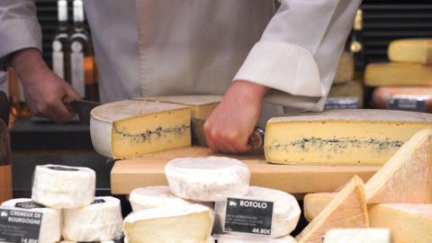 French cheese, white and yellow in different shapes and sizes, is displayed for sale at a supermarket. A man wearing a long white shirt is cutting a semi-circle of cheese in half, you can only see his arms and torso.