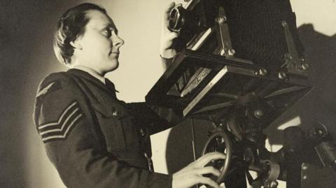 Sepia portrait of WAAF Sergeant Dorothy "Knicky" Chapman, standing in front of a large camera at RAF Iver Heath (Pinewood Studios). She is wearing her military uniform with sergeant's stripes on the sleeve.