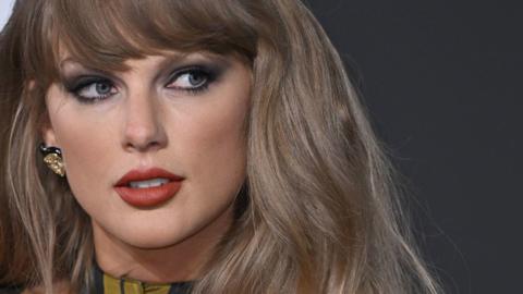 Close up of pop star Taylor Swift. She has dark blonde hair and is looking to the side. She has eyeliner on and a berry colour lipstick.