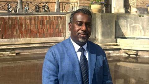 Yassin Mohamud wearing a white collared shirt and navy striped tie underneath a blue blazer. He is standing by still water outside Bristol City Council's offices on a sunny day.
