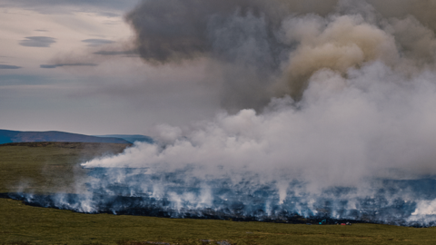 Aerial view of wide swathe of fire on charred moorland as smoke rises