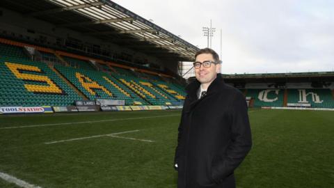 A man with glasses wearing a white shirt and black tie and black coat. He is standing with his hands in his pockets on the Franklins Garden's pitch. Two stands with green and yellow seats are in view behind him.