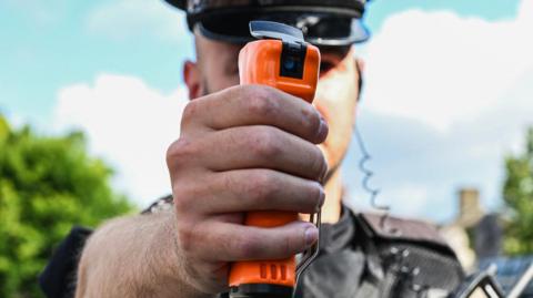 A police officer man holding an orange spray device in his hand - the device looks like a futuristic deodorant can.