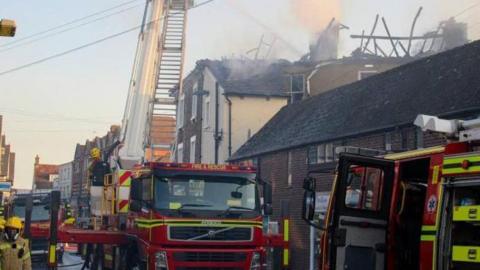 Fire engines and firefighters in a road of shops and flats above with smoke seen coming from the roof of a building.