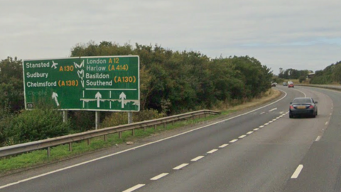 A street view image of the A12 with a black car approaching junction 19 at Chelmsford. On the left of the image there is a large green sign which says Stansted, Sudbury, Chelmsford, London, Harlow, Basildon, Southend, with arrows pointing either straight ahead or slightly to the left.