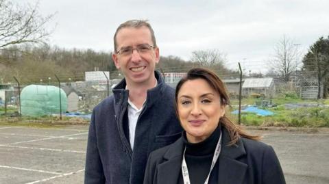 A man and a woman standing on an empty car park with allotments behind a fence in the background. The man is wearing a blue coat and a white shirt. He has short brown hair and has glasses. The woman has dark brown hair and is wearing a black top and coat and has a lanyard around her neck.