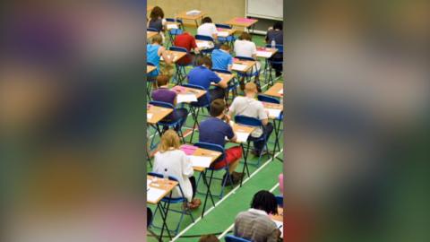 Young people sat at square desks in an exam hall. They are wearing a variety of colours and the floor is bright green.