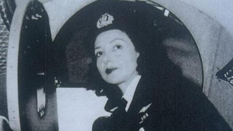 A black and white photo of Jackie Moggridge. She is wearing RAF uniform and is looking directly at the camera. She appears to be stood outside the cockpit of an aircraft. 