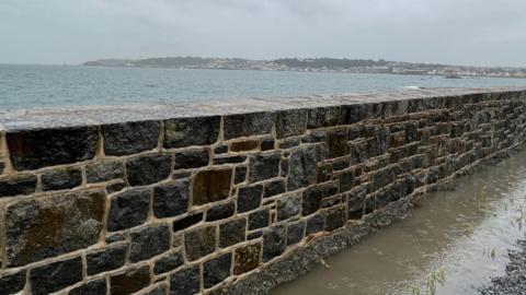 Rainfall in Guernsey - A modern brick wall taken at an angle with the floor flooded in brown water, behind the wall is the ocean with the island seen in the distance, raining with grey skies