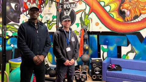 Two men, standing in a room, with graffiti on the walls, with a blue sofa to the right. They are both wearing caps and dark clothing and are smiling.
