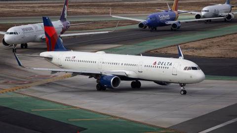 Two Delta airplanes sit on an airport tarmac with a line of planes behind them as thought they are waiting to take off  