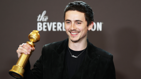 Timothee Chalamet in a black suit and black T-shirt holding up his Golden Globes award and smiling