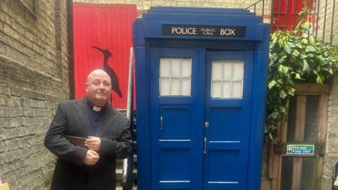Reverend Jason de-Vaux is standing on the left next to a replica of the Doctor Who Tardis which is blue and has a door on the right. It is sitting in a small courtyard.