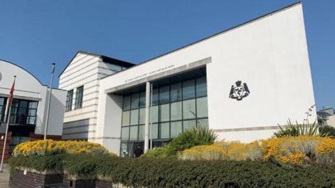 The Isle of Man Courts of Justice, which is a large white glass-fronted building with the government crest on the right hand side. There are bushed in planters in front of the building.