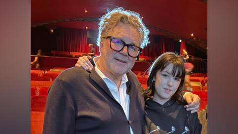 Fillm director Kirk Jones, left, an older man with a blue top and grey hair with glasses, with his arms around the shoulder of Megan Sykes, a younger woman with a black hoodie on and black hair. They are standing in front of several rows of red cinema seats