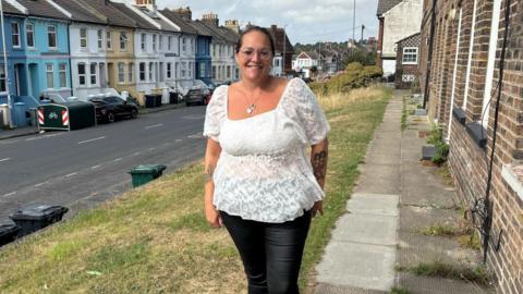 Kelly Warwick standing outside her house in Brighton. She wears a white blouse and dark jeans. She has dark hair tied up, and glasses.