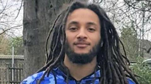 Nimroy Hendricks, who has a short beard and long dreadlocked hair and is wearing a tracksuit top with a blue camouflage design. He is stood in front of a tree, looking gently at the camera.