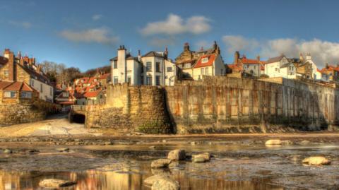 General view of Robin Hood's Bay, with historic houses above a high sea wall, and a rocky beach in the foreground.
