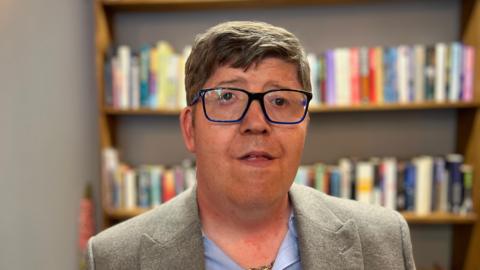 A man in his early forties with glasses and an eye condition looking towards the camera.