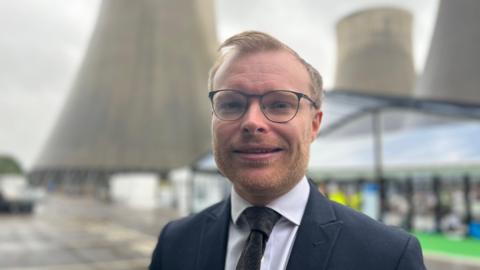 A ginger haired man wearing a suit and glasses stands in front of cooling towers.