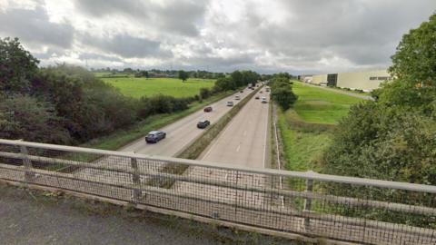 A view from a bridge of a dual carriageway running below and into the distance. Fields and trees are either side of the road. A large factory building is on the right. 