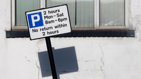 A white parking sign with a blue boxed in P, and a 2 hour limit. A white 