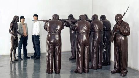 Two men in black caps stand next to a bronze figure of Jesus Christ, who is being held at rifle-point by six identical bronze figures in a semi-circular formation. A seventh bronze figure stand behind them, also holding a rifle, and resembling Mao Zedong