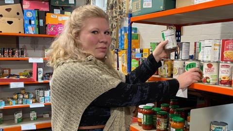 Becky Marshall, a middle-aged woman with blonde hair, stacking tins of soup on a full shelf of food supplies.