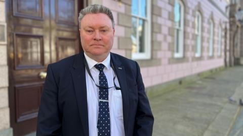 Jersey education minister Deputy Rob Ward stands outside a government building which has a large brown wooden door and lilac coloured brick work. He is wearing a dark suit and a white shirt with a black and white polka dot tie.