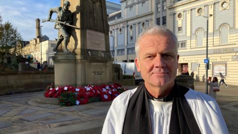 A man in black and white vicar's robes standing in front a war memorial with a stone statue of a soldier