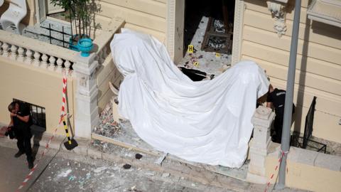 Frontage of a large cream coloured residential building in Monaco with damage to windows and a white sheet across the front steps and police standing near the front of the building. Photo: 30 June 2026
